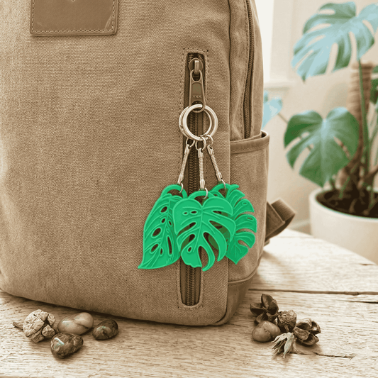 Green leaf-shaped keychain attached to a beige backpack with plants in the background