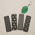 Four black metal bookmarks with geometric patterns and a green leaf-shaped keychain on a beige background.