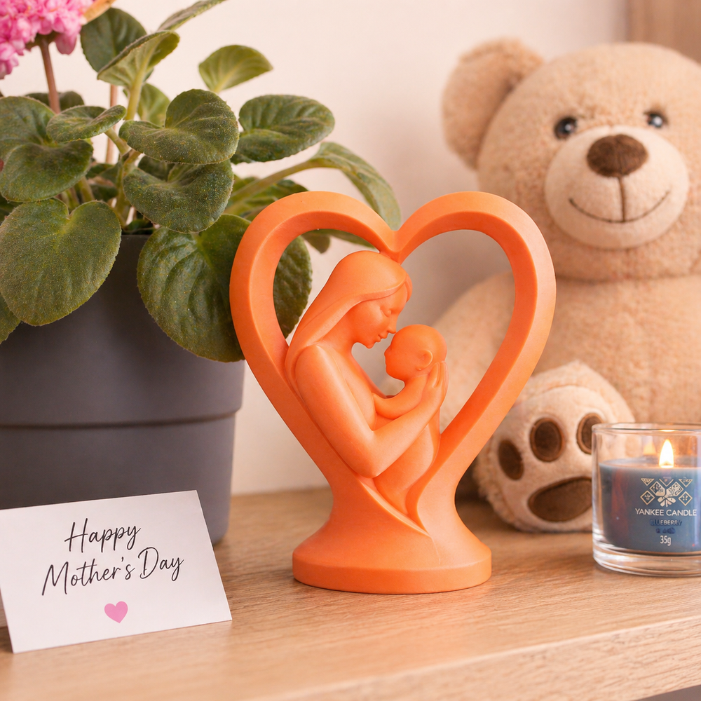 Orange sculpture of a mother holding a child with a heart-shaped frame, next to a potted plant, teddy bear, and candle.