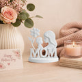 Decorative figurine with 'MOM' and a baby, surrounded by flowers and books on a table.