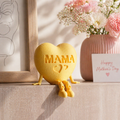 Yellow heart-shaped plush toy with 'MAMA' on it, sitting on a surface with flowers and a card in the background.