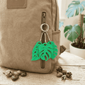 Green leaf-shaped keychain attached to a beige backpack with plants in the background