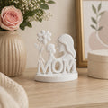 White decorative figurine with 'MOM' and a flower on a wooden surface with flowers and a vase in the background.