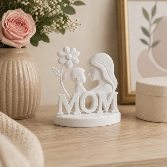 White decorative figurine with 'MOM' and a flower on a wooden surface with flowers and a vase in the background.